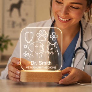 Può includere: Un'insegna luminosa a LED quadrata in acrilico con base in legno, caratterizzata da disegni al tratto incisi di un cane, un gatto, uno stetoscopio, un'impronta di zampa e una croce medica. Il testo Dr. Smith VETERINARY MEDICINE è illuminato.
