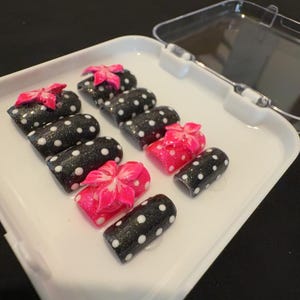Puede incluir: Un conjunto de uñas postizas negras y blancas con lunares y lazos rosas. Las uñas están dispuestas en una bandeja blanca, algunas con un acabado brillante. El tema general es el arte de uñas.