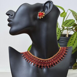 Könnte beinhalten: Ein rotes und goldenes Perlenkette- und Ohrring-Set, präsentiert auf einem schwarzen Büstenständer. Die Halskette hat ein detailliertes, kompliziertes Design. Die Ohrringe sind klein, rund und haben ein sternförmiges Design. Das Schmuckset ist auf einem schwarzen Büstenständer ausgestellt.