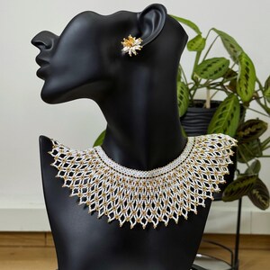 Puede incluir: Un collar de cuentas blanco y dorado y pendientes a juego sobre una cabeza de maniquí negra. El collar presenta un diseño geométrico. Los pendientes son pequeños, con forma de flor, dorados y blancos. La joya se exhibe sobre un fondo neutro.