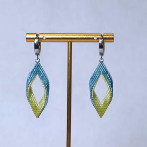 May include: A pair of beaded earrings with a teardrop shape. The earrings are blue and yellow, with a twisted design. They hang from silver-colored hoops and are displayed on a gold-colored earring stand.