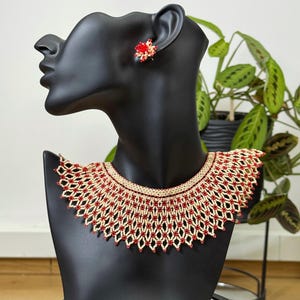 Puede incluir: Un collar de cuentas y pendientes a juego, exhibidos en un maniquí negro. El collar presenta una base color crema con detalles de cuentas rojas. Los pendientes son pequeños, rojos, con forma de flor. Las joyas se muestran sobre un fondo de plantas verdes.