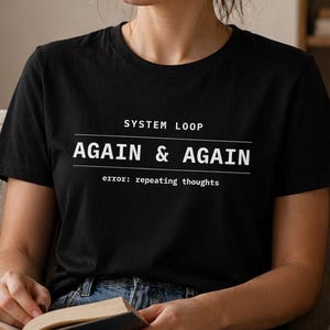 May include: Black t-shirt with the text "SYSTEM LOOP AGAIN & AGAIN error: repeating thoughts" in white. The shirt is short-sleeved and worn by a person sitting on a beige couch, holding a book.