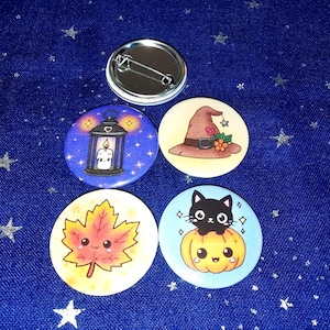 Könnte beinhalten: Ein Set aus vier runden Ansteckbuttons mit farbenfrohen Illustrationen. Ein Button zeigt eine Laterne mit Kerze, einer einen braunen Hexenhut, einer ein lächelndes Ahornblatt und einer eine schwarze Katze auf einem Kürbis. Ein Button ist von hinten zu sehen.