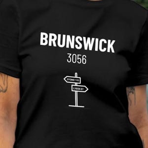 May include: A black short-sleeved t-shirt featuring white text that reads BRUNSWICK 3056 above a graphic of a street sign post with two directional signs labeled SYDNEY RD and LYGON ST. The shirt has a crew neckline and a standard fit.