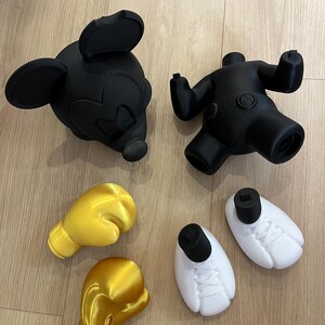 Puede incluir: Cabeza y cuerpo negros de Mickey Mouse, con guantes de boxeo amarillos y zapatos blancos. Las piezas est&aacute;n impresas en 3D y parecen ser parte de un conjunto de esculturas o juguetes m&aacute;s grande. Los art&iacute;culos est&aacute;n dispuestos sobre una superficie de madera clara.