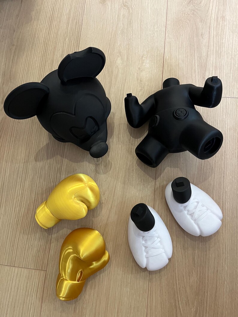 Puede incluir: Cabeza y cuerpo negros de Mickey Mouse, con guantes de boxeo amarillos y zapatos blancos. Las piezas est&aacute;n impresas en 3D y parecen ser parte de un conjunto de esculturas o juguetes m&aacute;s grande. Los art&iacute;culos est&aacute;n dispuestos sobre una superficie de madera clara.