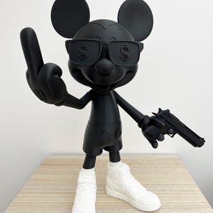 Könnte beinhalten: Schwarze Mickey Mouse-Figur mit Sonnenbrille mit Dollarzeichen, die eine Pistole hält und den Mittelfinger zeigt. Die Figur trägt weiße Turnschuhe und schwarze Shorts.