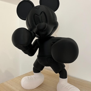 Puede incluir: Figura negra de Mickey Mouse en pose de boxeo, con guantes y pantalones cortos negros y zapatos blancos. La figura tiene una mordida en una oreja. La figura est&aacute; sobre una superficie marr&oacute;n claro.