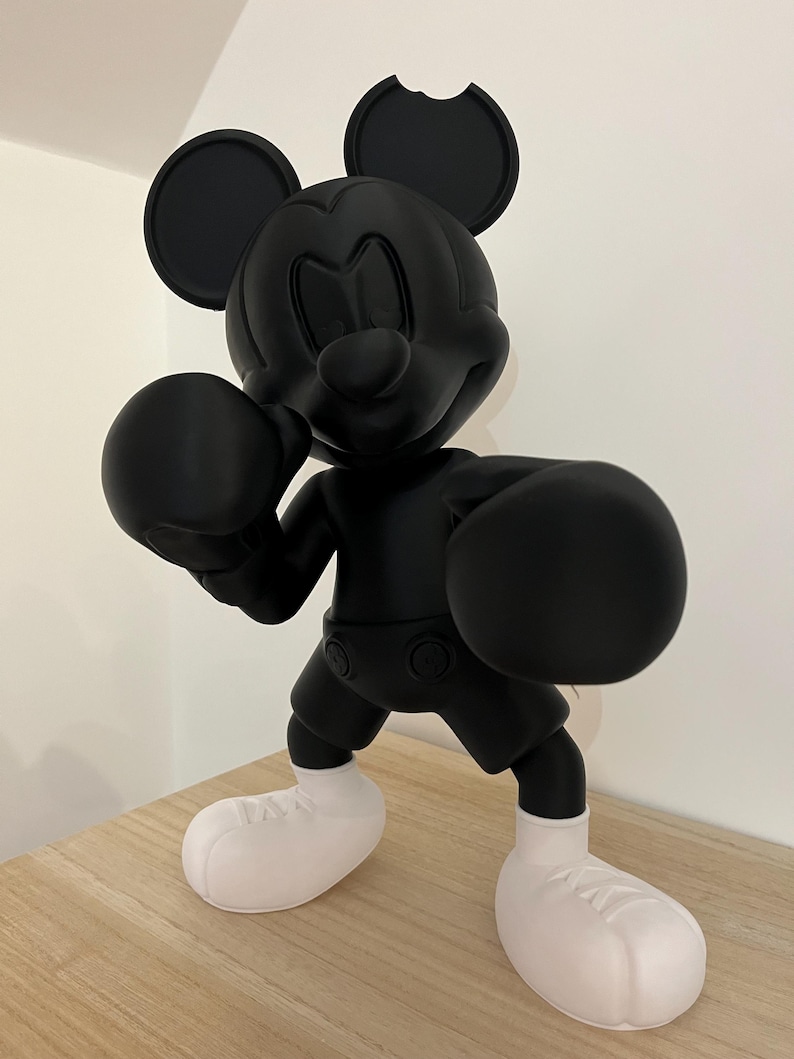 Puede incluir: Figura negra de Mickey Mouse en pose de boxeo, con guantes y pantalones cortos negros y zapatos blancos. La figura tiene una mordida en una oreja. La figura est&aacute; sobre una superficie marr&oacute;n claro.