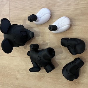 Puede incluir: Colecci&oacute;n de objetos impresos en 3D en blanco y negro, que incluyen una cabeza de Mickey Mouse, dos guantes de boxeo, dos objetos blancos en forma de huevo con accesorios cil&iacute;ndricos negros y un objeto negro similar a una c&aacute;mara. Dispuestos sobre una superficie de madera marr&oacute;n claro.