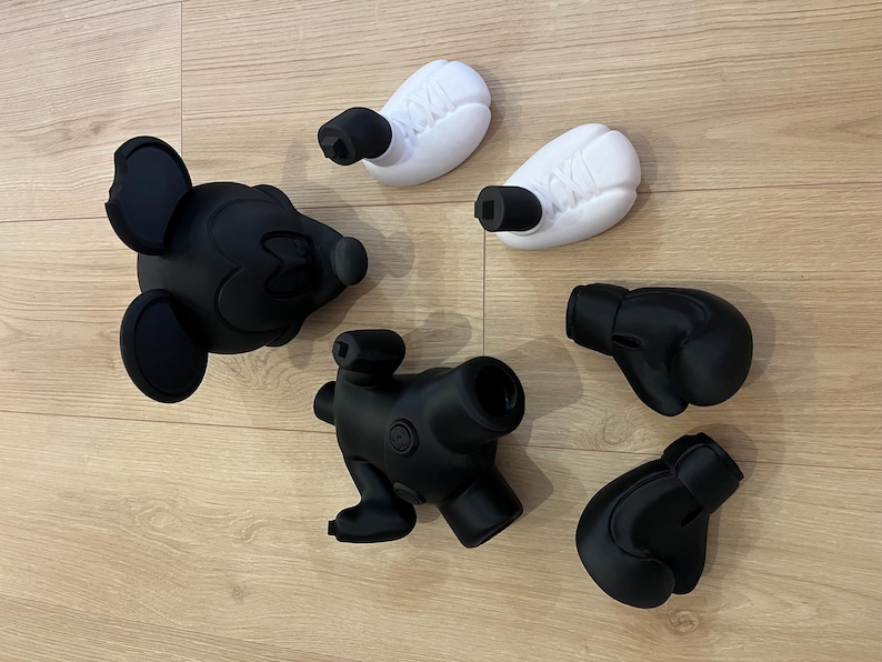 Puede incluir: Colecci&oacute;n de objetos impresos en 3D en blanco y negro, que incluyen una cabeza de Mickey Mouse, dos guantes de boxeo, dos objetos blancos en forma de huevo con accesorios cil&iacute;ndricos negros y un objeto negro similar a una c&aacute;mara. Dispuestos sobre una superficie de madera marr&oacute;n claro.