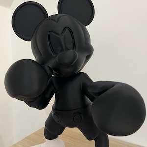 Puede incluir: Una figura negra de Mickey Mouse en posici&oacute;n de boxeo, con grandes guantes de boxeo negros y zapatos blancos. La figura tiene un acabado mate y est&aacute; posada con los brazos en alto, lista para pelear. Las orejas son redondas, y una oreja tiene una mordida.