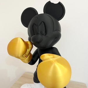 Puede incluir: Figura negra de Mickey Mouse en pose de boxeo, con guantes y pantalones cortos de boxeo dorados. La figura tiene zapatos blancos y una mordida en una oreja. La figura est&aacute; sobre una superficie marr&oacute;n claro.