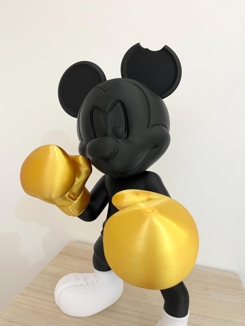 Puede incluir: Figura negra de Mickey Mouse en pose de boxeo, con guantes y pantalones cortos de boxeo dorados. La figura tiene zapatos blancos y una mordida en una oreja. La figura est&aacute; sobre una superficie marr&oacute;n claro.