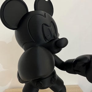 Puede incluir: Figura negra de Mickey Mouse en pose de boxeo, con pantalones cortos, guantes y zapatos negros con suelas blancas. Una oreja est&aacute; parcialmente mordida. La figura est&aacute; sobre una superficie de madera.