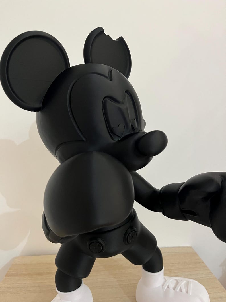 Puede incluir: Figura negra de Mickey Mouse en pose de boxeo, con pantalones cortos, guantes y zapatos negros con suelas blancas. Una oreja est&aacute; parcialmente mordida. La figura est&aacute; sobre una superficie de madera.