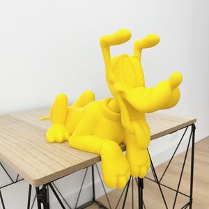 Peut inclure: Une sculpture jaune vif d'un chien de dessin animé, allongé avec la langue sortie. Le chien est posé sur une petite table en bois clair avec un cadre en métal noir. L'arrière-plan est un mur blanc uni.