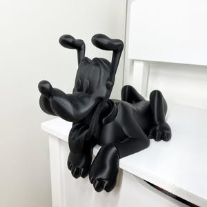 Pode incluir: Escultura preta de Pluto, personagem da Disney, deitado. As orelhas estão levantadas. A figura está colocada sobre uma superfície branca, com um fundo branco.