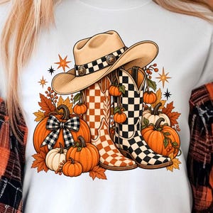 Puede incluir: Una camiseta blanca con un estampado gráfico de un sombrero de vaquero beige, dos botas vaqueras con patrones de cuadros y varias calabazas naranjas. El diseño incluye hojas de otoño, bayas pequeñas y estrellas rodeando los elementos.