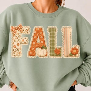Könnte beinhalten: Ein salbeigrünes Sweatshirt mit Rundhalsausschnitt und dem Wort FALL in großen, applizierten Buchstaben. Jeder Buchstabe ist aus verschiedenen gemusterten Stoffen mit Spitzenbesatz gefertigt, darunter florale, karierte und gestreifte Designs.