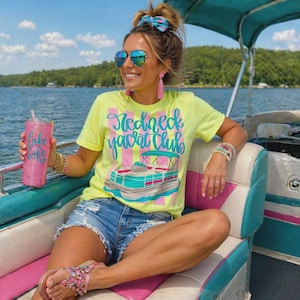 May include: A woman wearing a neon yellow t-shirt with the words "Redneck Yacht Club" and a boat graphic. She is wearing blue jean shorts, sunglasses, and holding a pink glitter tumbler that says "Lake Life".