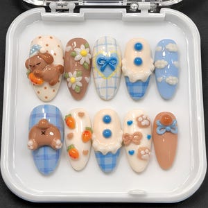 Puede incluir: Un conjunto de diez uñas postizas con varios diseños. Las uñas presentan una gama de colores, incluyendo marrón, azul y blanco, con elementos decorativos como lazos, flores y personajes de dibujos animados. Algunas uñas tienen un patrón de cuadros, otras un color sólido.