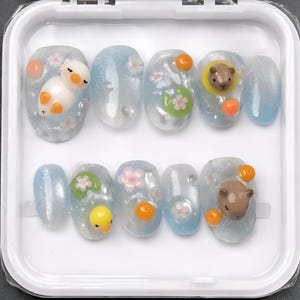May include: A set of ten artificial nails in a clear plastic case. The nails are light blue with glitter and feature 3D designs of ducks, bears, flowers, and orange accents. The nails are oval-shaped.