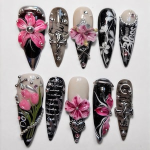 Puede incluir: Una colección de diez uñas artificiales largas y puntiagudas. Cada uña presenta diseños únicos, que incluyen acentos florales en 3D, adornos metálicos y patrones intrincados. Los colores incluyen negro, rosa, beige y plateado. Algunas uñas tienen texto.