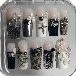 May include: A set of ten artificial nails in a clear plastic case. The nails feature a variety of designs, including black lace, silver charms, leopard print, and the word "Angel". Some nails have a nude base with embellishments.