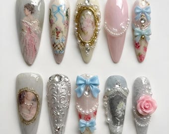 Puede incluir: Una colección de diez uñas artificiales largas en forma de almendra. Cada uña presenta diseños intrincados, incluyendo retratos, lazos, perlas y patrones florales. Los colores incluyen rosa, blanco, azul y dorado, con un acabado brillante.
