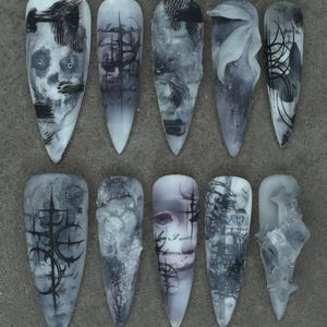 Puede incluir: Un conjunto de diez uñas postizas largas y puntiagudas con diseños góticos. Las uñas presentan obras de arte oscuras en escala de grises, incluyendo calaveras, patrones abstractos y elementos florales. Cada uña tiene un diseño único.