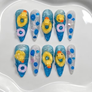 Puede incluir: Un conjunto de diez uñas artificiales largas y puntiagudas. Cada uña presenta una base azul con un patito de goma amarillo y un flotador rosa y blanco. Algunas uñas tienen detalles azules y plateados.