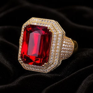 Peut inclure: Une bague dorée ornée d'une grande pierre rouge rectangulaire. La pierre est entourée d'un double halo de petites pierres claires. Le corps de la bague est également incrusté de petites pierres, créant un aspect luxueux.