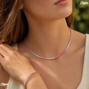 May include: A tennis necklace and matching tennis bracelet featuring a gradient of round-cut stones that transition in color from deep pink to light pink and clear white. The jewelry pieces are set in a metallic frame and worn around the neck.