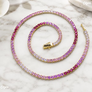 May include: A gold-tone tennis necklace featuring a continuous row of round-cut stones in a gradient color scheme, transitioning from deep magenta and vibrant pink to light lavender and white, secured with a rectangular box clasp closure.