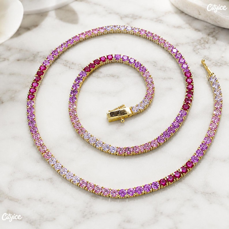 May include: A gold-tone tennis necklace featuring a continuous row of round-cut stones in a gradient color scheme, transitioning from deep magenta and vibrant pink to light lavender and white, secured with a rectangular box clasp closure.
