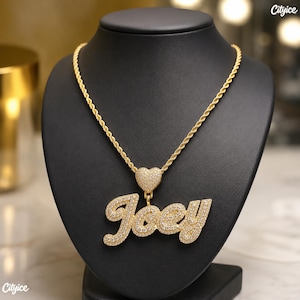 May include: A gold-tone rope chain necklace featuring a pendant with the name Joey in cursive script, encrusted with small clear stones. A small heart-shaped charm, also covered in clear stones, connects the pendant to the chain.
