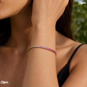 May include: A gold-toned tennis bracelet featuring a continuous row of round-cut stones that transition in color from clear white to light pink and finally to a deep, vibrant magenta, secured with a metallic clasp on a wrist.