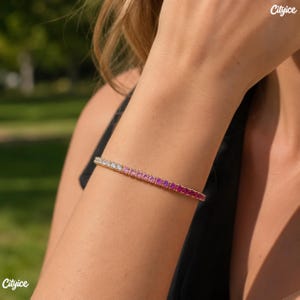 May include: A gold-toned tennis bracelet featuring a gradient of round-cut stones that transition from clear white to light pink and deep magenta. The stones are set in a continuous line, creating a vibrant, multi-colored effect on the wrist.