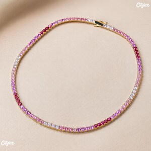 May include: A gold-tone tennis necklace featuring a continuous row of round-cut stones in a gradient of colors, transitioning from deep red and magenta to light pink and clear white, secured with a metal box clasp closure at the top.