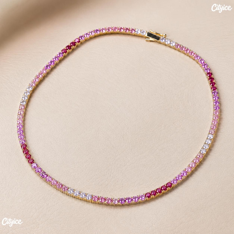 May include: A gold-tone tennis necklace featuring a continuous row of round-cut stones in a gradient of colors, transitioning from deep red and magenta to light pink and clear white, secured with a metal box clasp closure at the top.