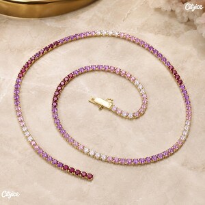 May include: A gold-tone tennis necklace featuring a continuous row of round-cut stones in a gradient color scheme, transitioning from deep red and magenta to light pink and white. The necklace includes a secure metal box clasp closure.