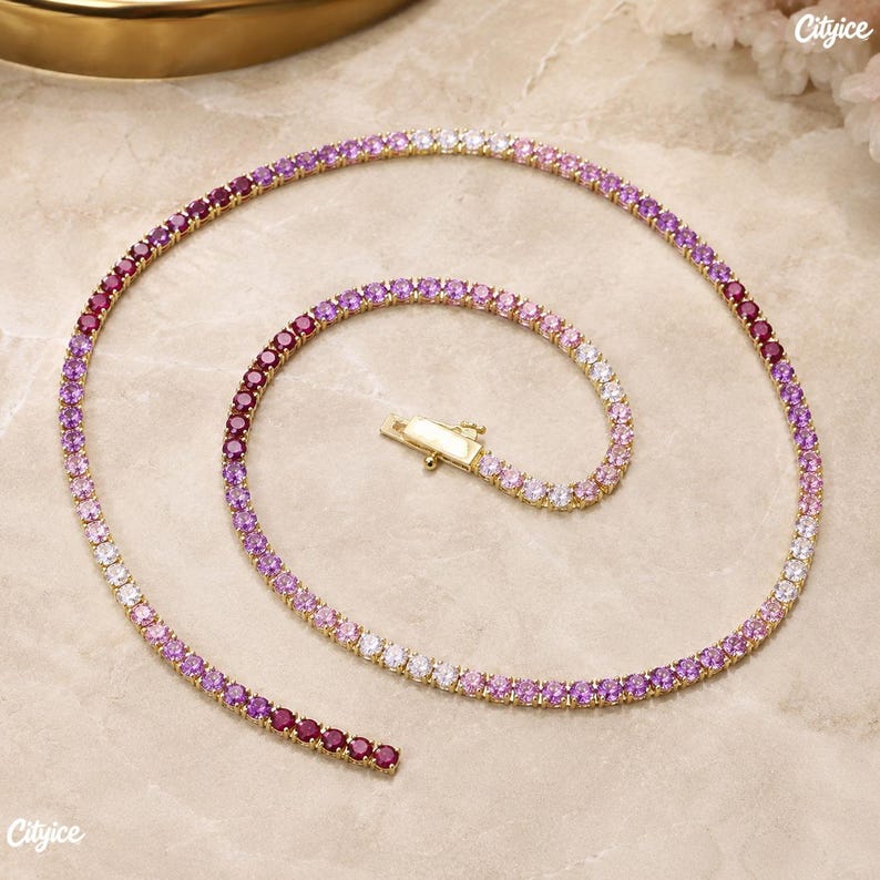 May include: A gold-tone tennis necklace featuring a continuous row of round-cut stones in a gradient color scheme, transitioning from deep red and magenta to light pink and white. The necklace includes a secure metal box clasp closure.