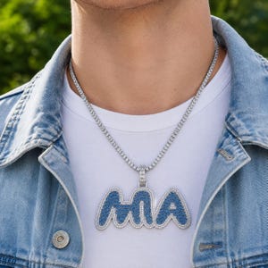 May include: A silver chain necklace with a pendant spelling out "MA" in a blue bubble letter design. The pendant is encrusted with small, clear stones. The necklace is worn over a white t-shirt and a denim jacket.