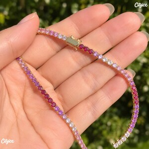 May include: A gold-tone tennis necklace featuring a gradient of round-cut stones transitioning from deep red and magenta to light pink and clear white. The necklace includes a secure rectangular box clasp closure with a side safety latch.