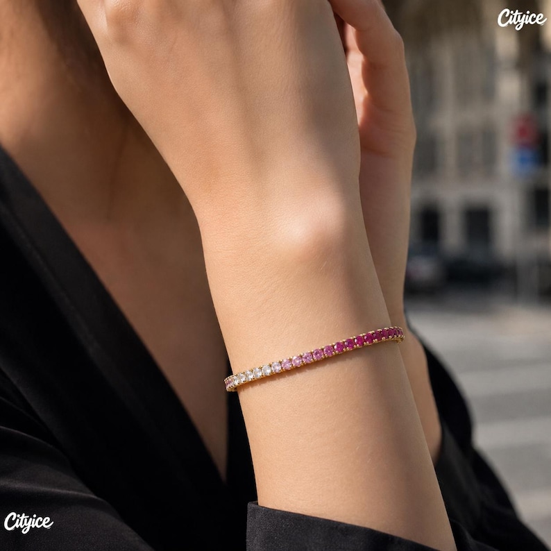 May include: A gold-toned tennis bracelet featuring a gradient of round-cut stones that transition in color from clear white to light pink and deep magenta. The stones are set in a continuous line, creating a vibrant and polished accessory.