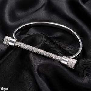 May include: A silver-toned metal bangle bracelet featuring a horizontal bar centrepiece. The bar is encrusted with numerous small, clear, sparkling stones, and the ends of the bar are capped with smooth, polished silver-toned metal cylinders.
