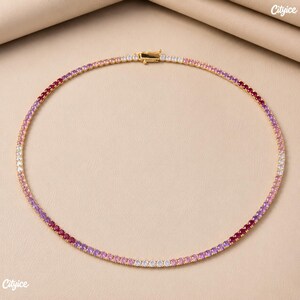 May include: A gold-tone tennis necklace featuring a continuous row of round-cut stones in a gradient color pattern. The stones transition through shades of deep red, magenta, light pink, and clear white. It includes a secure metal box clasp closure.