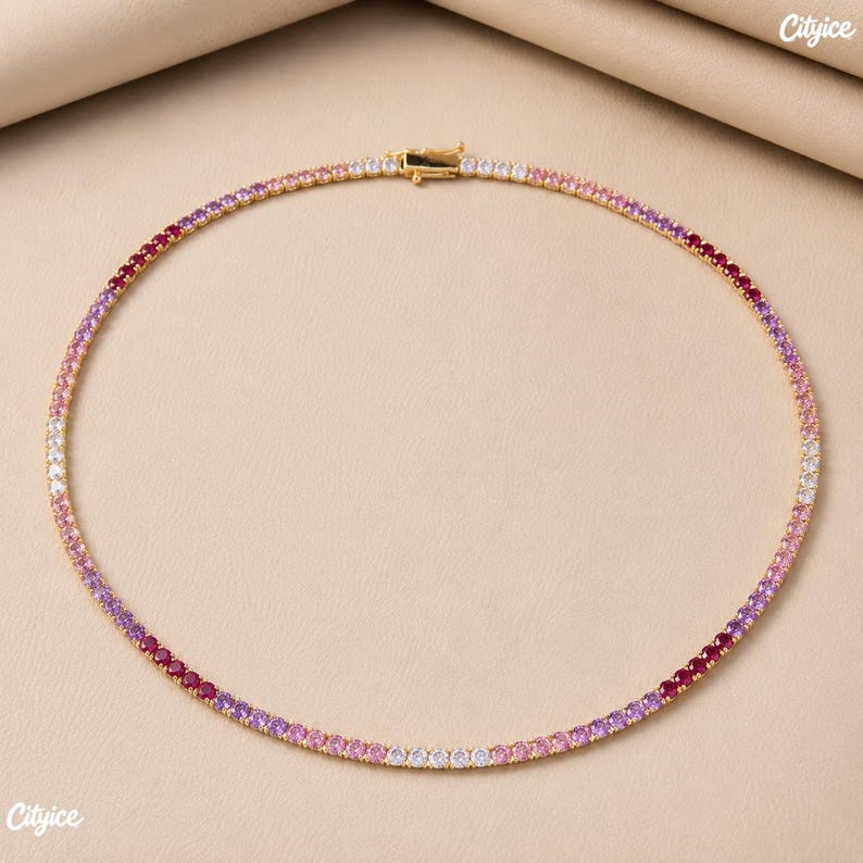 May include: A gold-tone tennis necklace featuring a continuous row of round-cut stones in a gradient color pattern. The stones transition through shades of deep red, magenta, light pink, and clear white. It includes a secure metal box clasp closure.